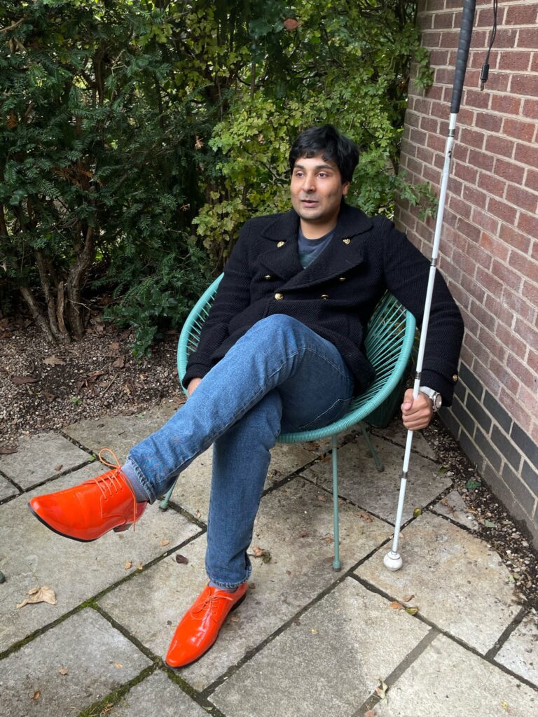 Kaleem sat outdoors in a black jacket, blue jeans and bright fluorescent orange shoes. Kaleem holds his white cane beside him in his left hand.